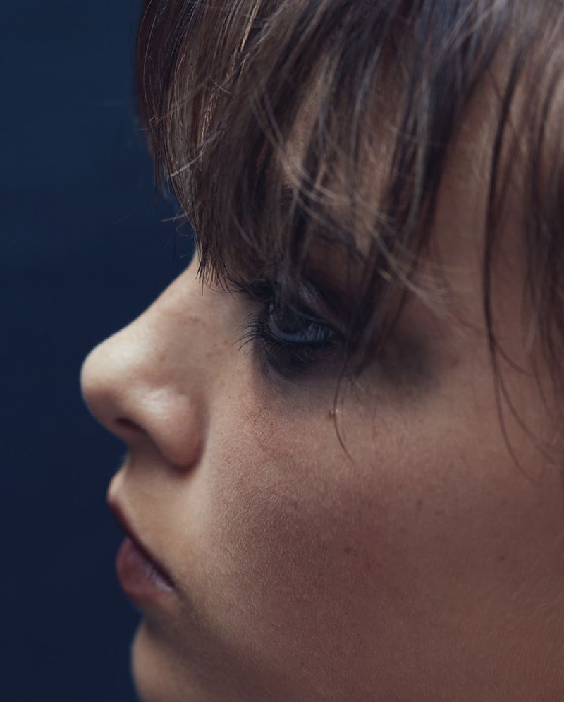Close-up profile of an adult female with brown hair and soft lighting against a dark blue background focusing on her facial features.