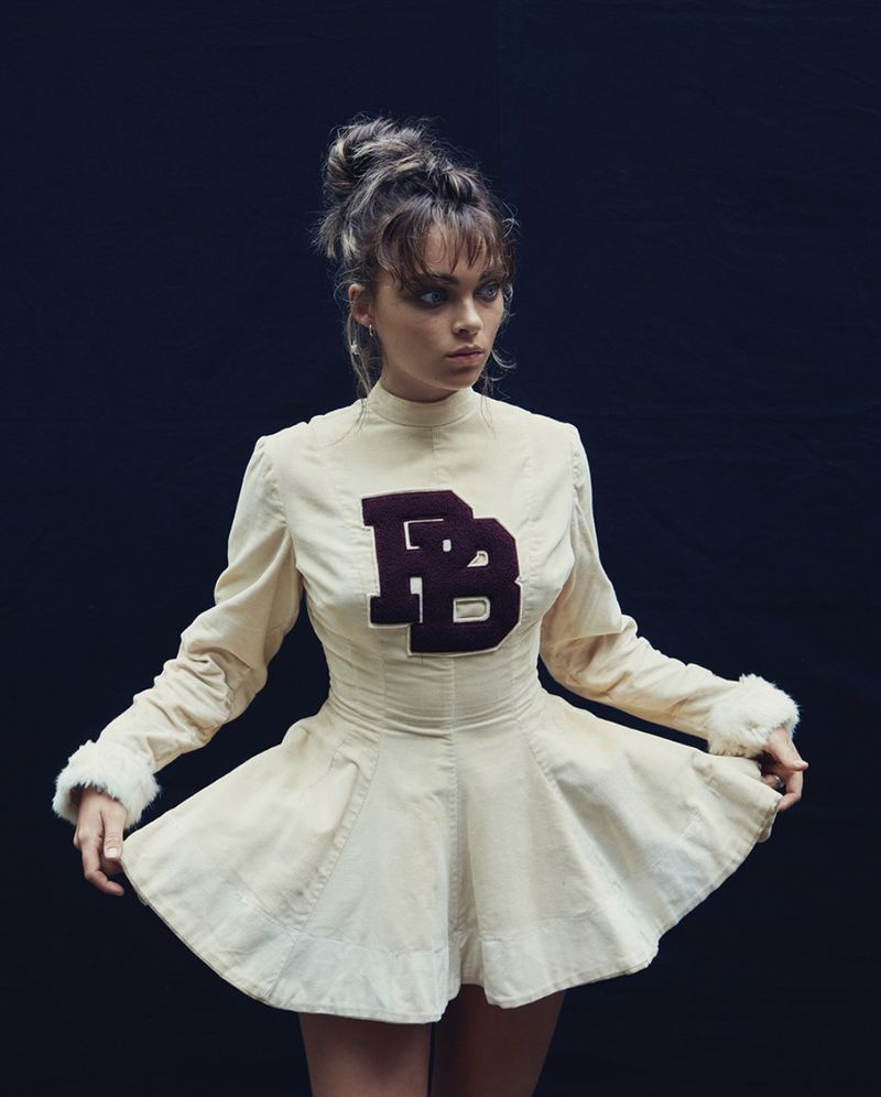 Portrait of a young woman in a cream-colored dress with a 'BB' logo, styled with a messy bun and dark eye makeup, against a dark backdrop.