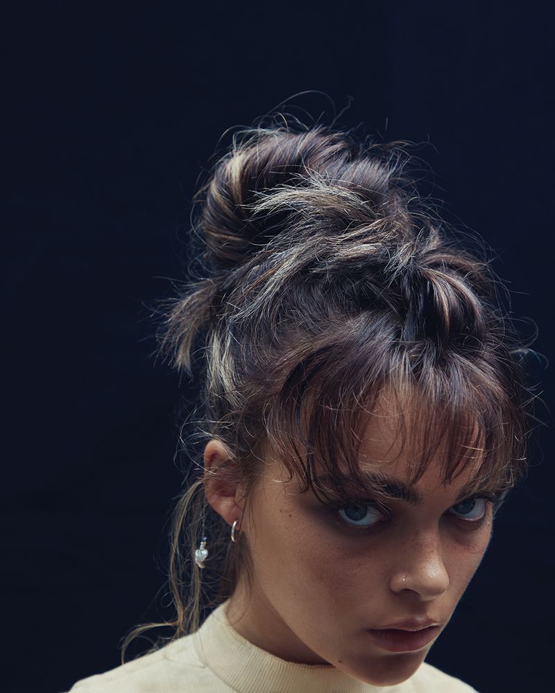 Close-up studio portrait of a young adult woman with dark hair, wearing a cream-colored top, and a silver hoop earring.