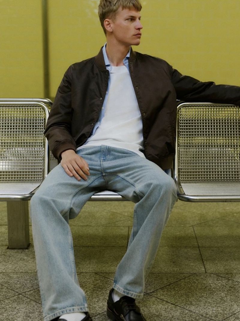 Model Timo Pan wearing a dark brown jacket, white t-shirt, and light wash denim jeans seated on a metal bench.