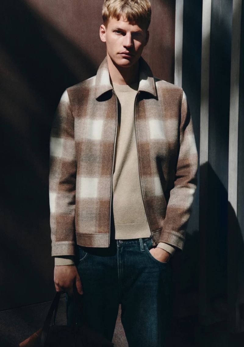 Model Timo Pan wearing a brown plaid zip jacket, beige knit sweater, and blue jeans against a shadowed backdrop.