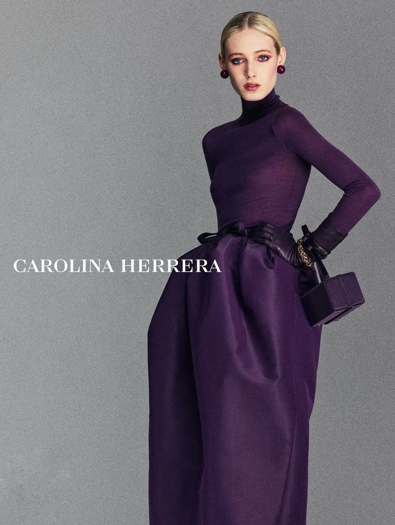 Model posing in a deep purple high-neck top, voluminous structured skirt, matching gloves, and a box bag against a grey backdrop.