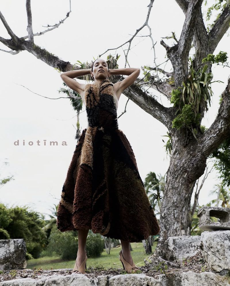 Model Romae Gordon in a textured brown midi dress posing with arms raised in front of an ancient tree outdoors.