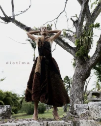 Model Romae Gordon in a textured brown midi dress posing with arms raised in front of an ancient tree outdoors.