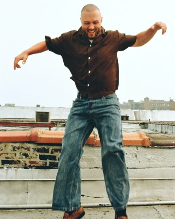 Music producer Chris Lake jumping mid-air on an urban rooftop wearing a brown button-down shirt and baggy blue jeans.