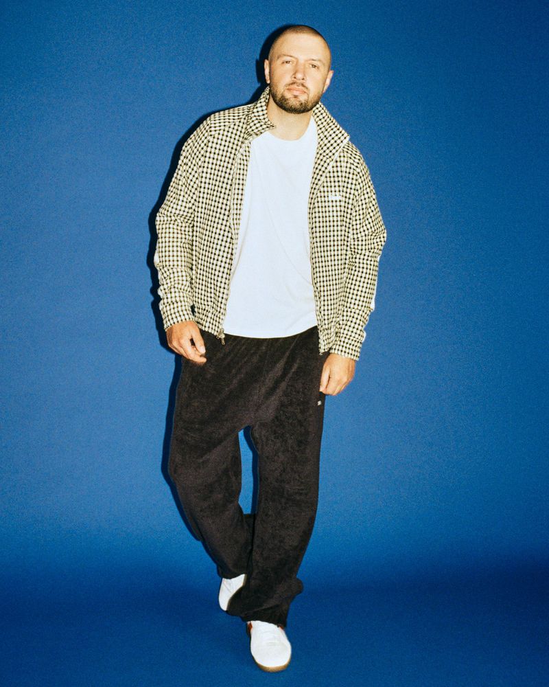 Chris Lake posing in a patterned jacket, black trousers, and white sneakers against a solid blue backdrop.