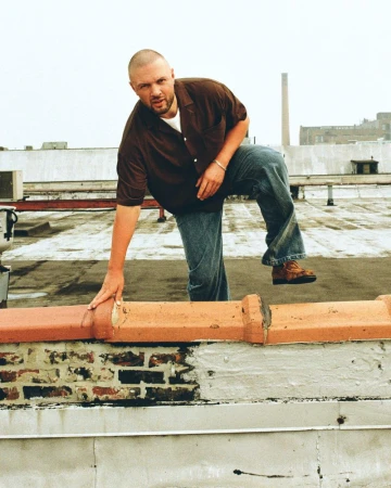 Chris Lake leaning over a brick rooftop parapet wearing a brown short-sleeve shirt and baggy blue denim jeans.