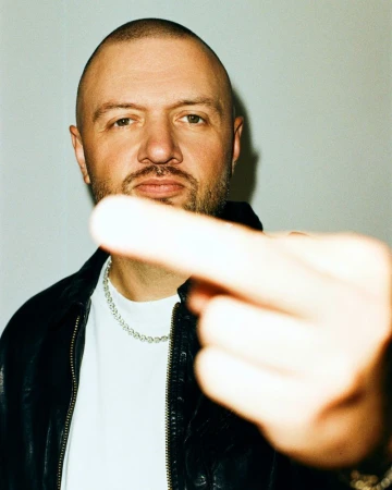 Close-up portrait of music producer Chris Lake gesturing toward the camera in a black leather jacket and silver chain.