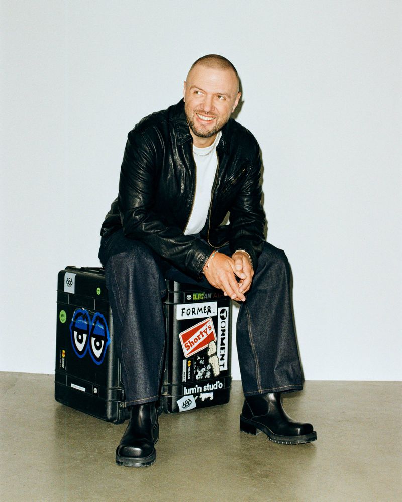 DJ Chris Lake sitting on a sticker-covered flight case wearing a black leather jacket and wide-leg denim against a white studio wall.