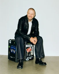Chris Lake smiling while seated on a sticker-covered black flight case wearing a black leather jacket and dark wide-leg denim jeans.