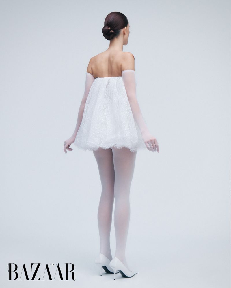 A woman models a strapless, white lace dress, sheer gloves, and white tights and heels in a studio with a white background.