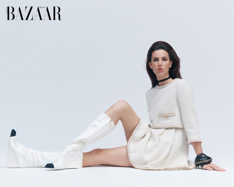 Elegant woman in a white tweed dress and white boots poses in a light studio. She has her dark hair down and wears a black choker.