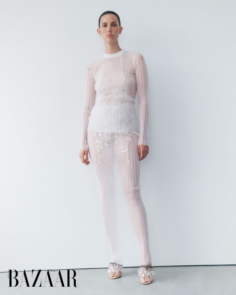 Elegant fashion shot featuring a female model in a sheer white, long-sleeved top and skirt with embellishments, posing against a plain white background.