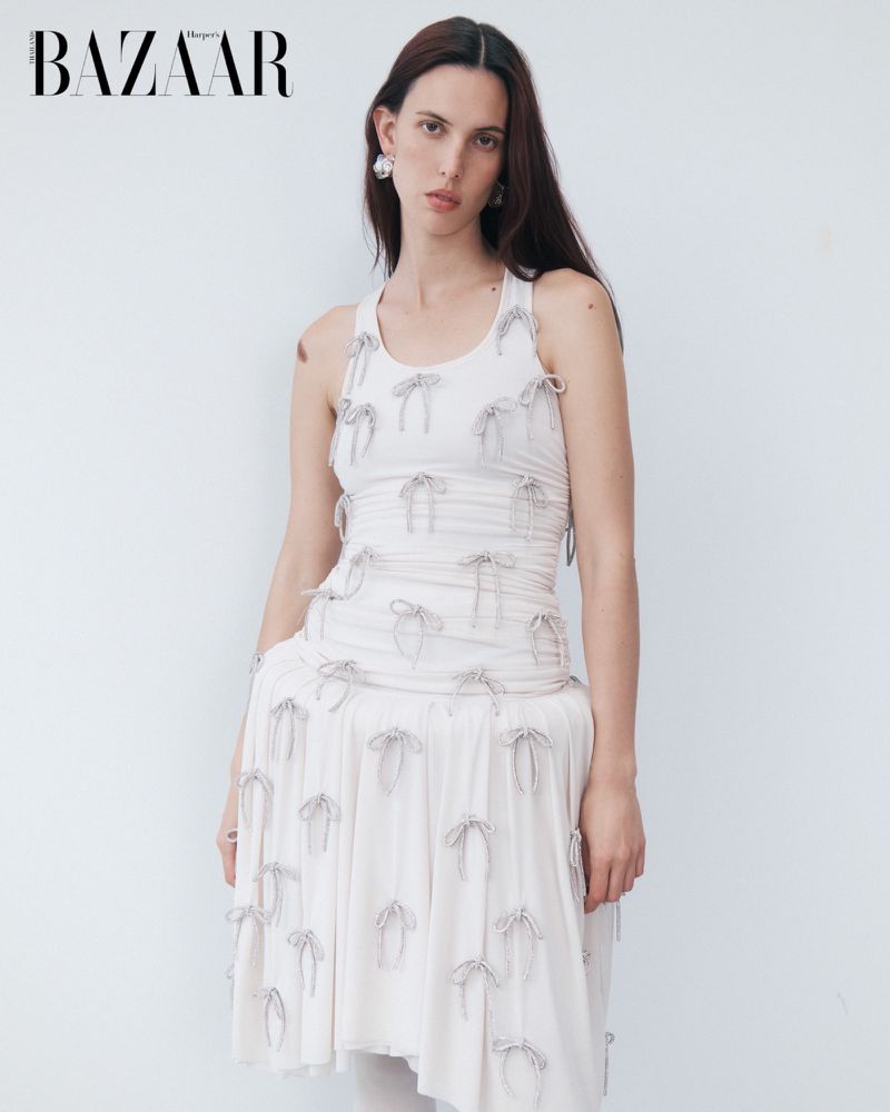 A young woman with long brown hair poses against a white wall wearing a white tank dress adorned with tiny decorative bows.