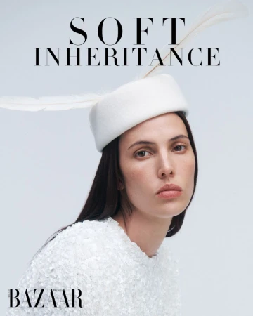 Portrait of an adult female fashion model in a white textured top and feathered hat against a light grey background in a studio.