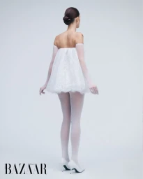 Model from behind in a white strapless mini dress, sheer gloves, tights, and heels against a pale grey seamless background.