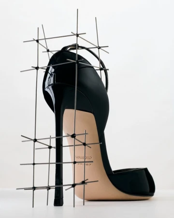 Black high-heeled stiletto pump enclosed in a delicate wire grid scaffold against a bright white background.