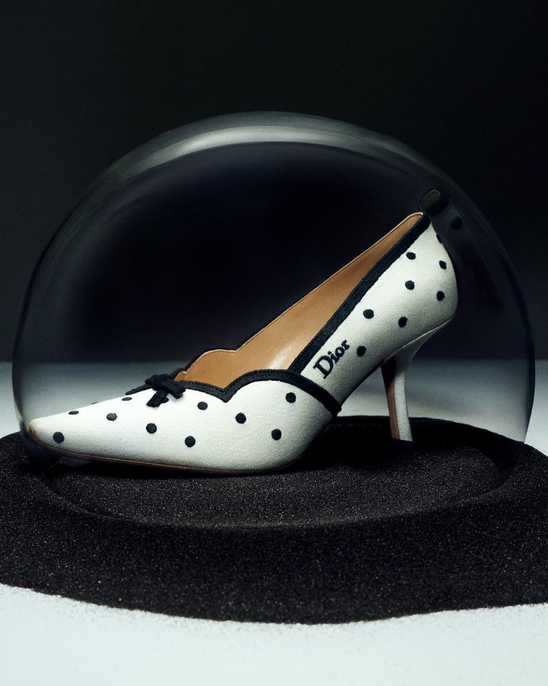 White pointed-toe pump with black polka dots and a Dior logo enclosed inside a clear glass cloche over black sand.