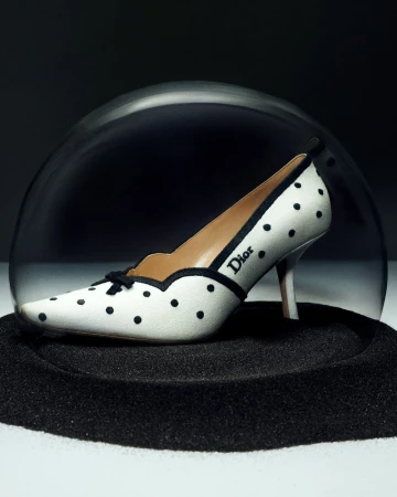 White pointed-toe pump with black polka dots and a Dior logo enclosed inside a clear glass cloche over black sand.