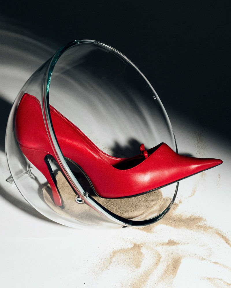 Red pointed-toe high heel pump resting inside a clear glass bowl with fine sand on a white surface with hard shadows.