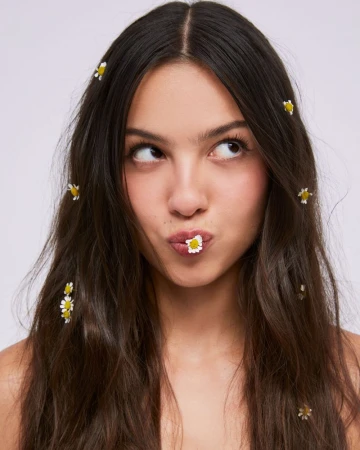 Olivia Rodrigo with daisies woven through her dark brown hair and a daisy in her mouth against a pale backdrop.