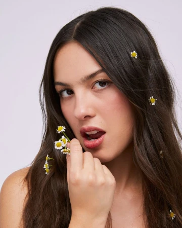 Portrait of Olivia Rodrigo holding tiny white daisy blossoms near her lips with flowers woven into her dark brown hair.