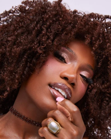 Close-up beauty portrait of a Black woman with voluminous curly brown hair wearing glossy lips, a beaded choker, and a statement ring.