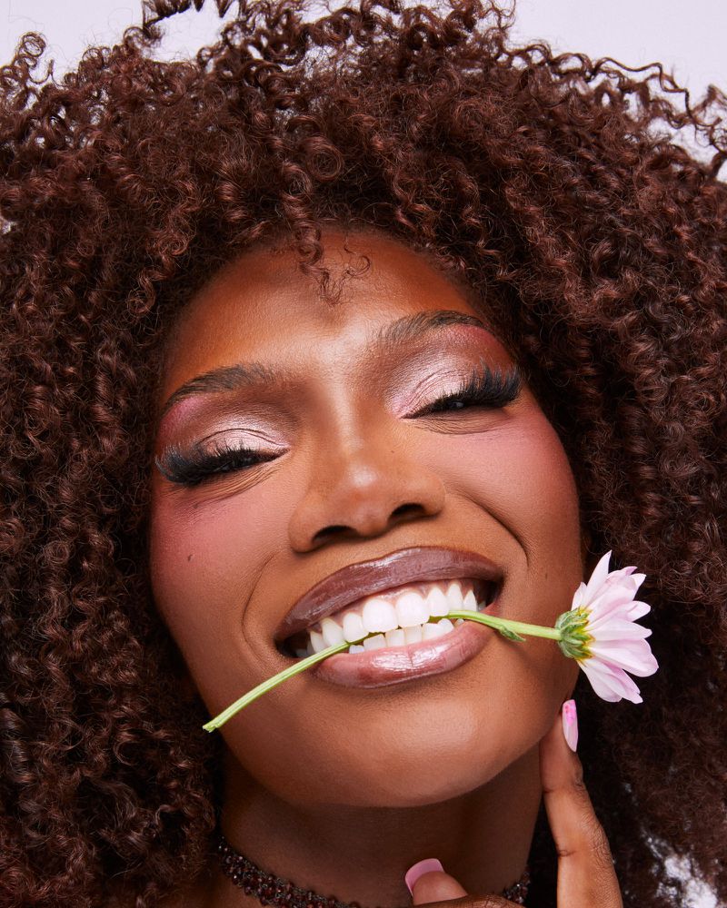 Close-up portrait of a smiling Black woman with voluminous curly hair holding a pink flower stem between her teeth.