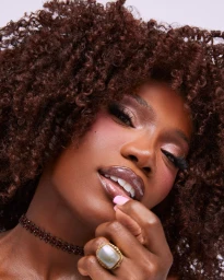 Close-up beauty portrait of a Black woman with voluminous curly brown hair, glossy lips, a beaded choker, and a large statement ring.