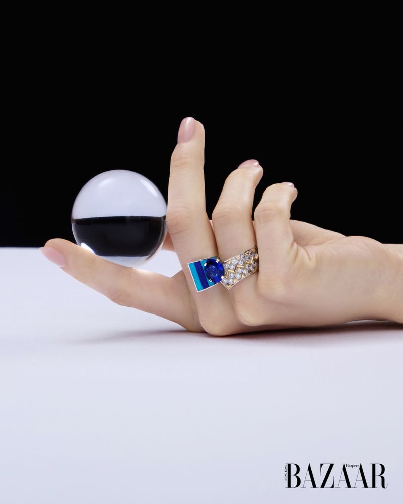 A hand wearing diamond and blue gemstone rings balances a clear glass sphere on a white surface against a black background.