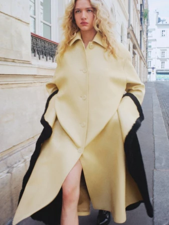 Model Mila Prvulovic walking along a city sidewalk wearing an oversized pale yellow coat with contrasting black lining.