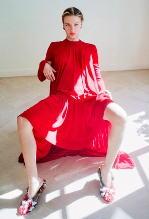 Model Mila Prvulovic seated wearing a sweeping vibrant red high-neck pleated dress and patterned mules in a sunlit room.