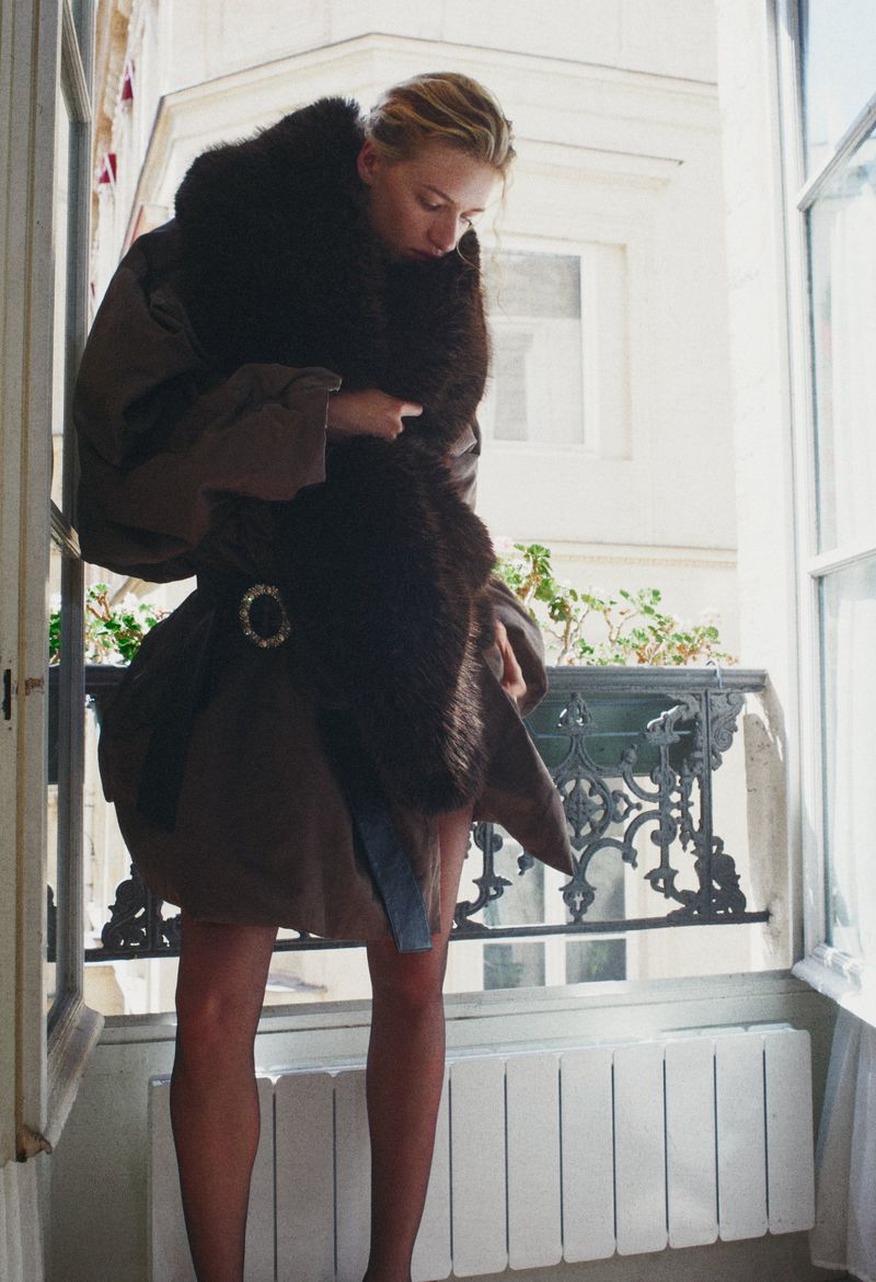 Model Mila Prvulovic wearing a brown belted coat and plush dark fur stole by an open window balcony.