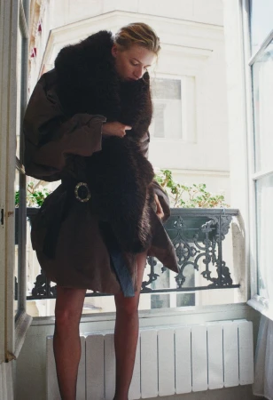 Model Mila Prvulovic wearing a brown belted coat and plush dark fur stole by an open window balcony.