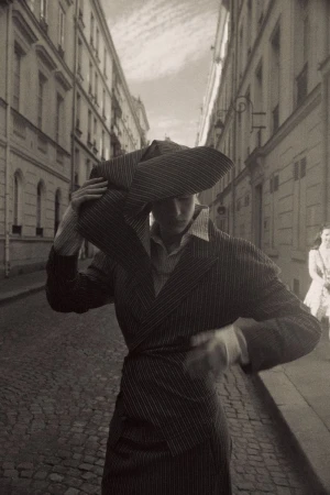 Model Mila Prvulovic wearing a structured pinstripe suit and sculptural hat on a European cobblestone street.