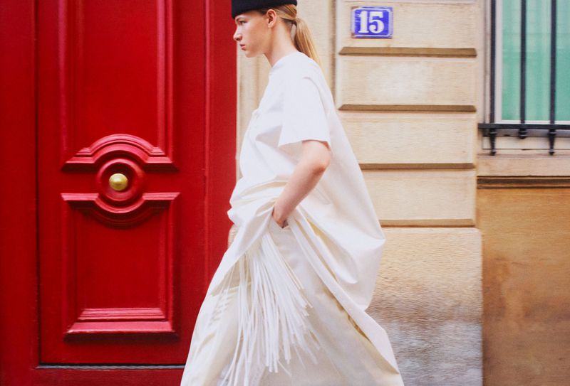 Model Mila Prvulovic walking in a white fringed dress past a vibrant red wooden door and building facade.