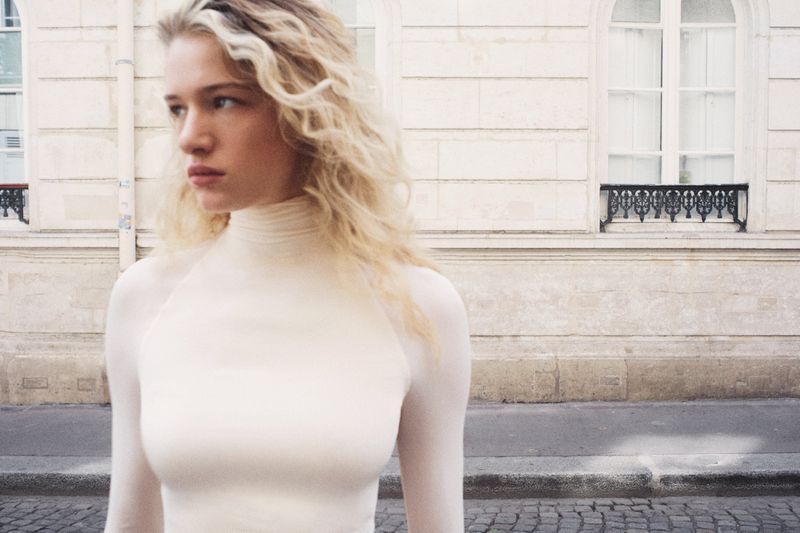 Mila Prvulovic wearing a cream long-sleeve turtleneck top standing on a street in front of a stone building facade.