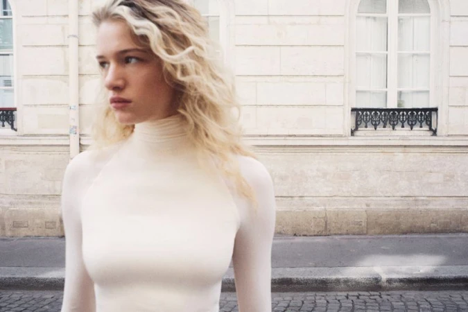 Mila Prvulovic wearing a cream long-sleeve turtleneck top standing on a street in front of a stone building facade.