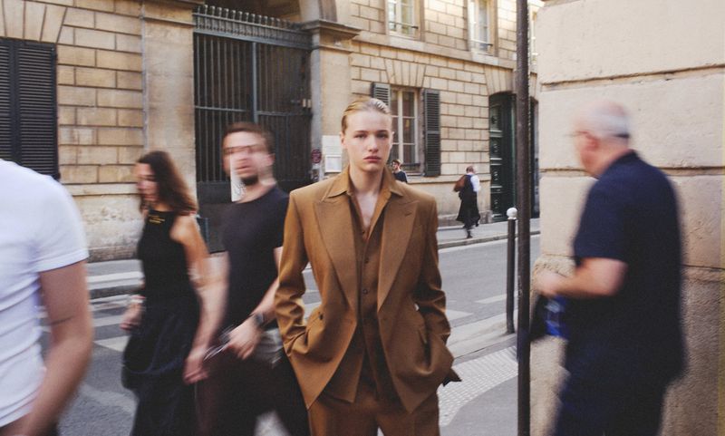 Model Mila Prvulovic stands in a tailored caramel brown suit on a bustling city street with blurred pedestrians walking past.