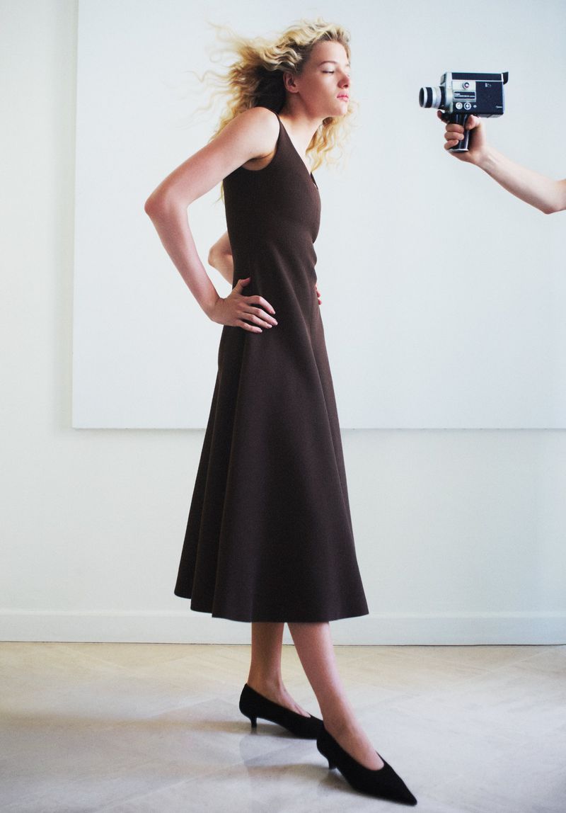 Model Mila Prvulovic standing in a sleeveless brown midi dress with hands on hips as a vintage handheld camera is pointed at her.