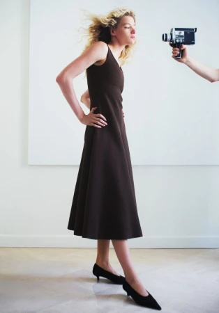 Model Mila Prvulovic standing in a sleeveless brown midi dress with hands on hips as a vintage handheld camera is pointed at her.