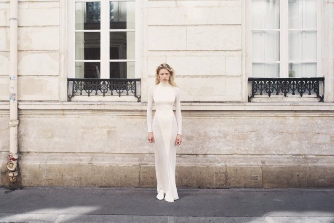 Model Mila Prvulovic in a floor-length white high-neck gown standing against a limestone building exterior with ornate window grilles.