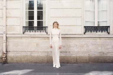 Model Mila Prvulovic in a floor-length white high-neck gown standing against a limestone building exterior with ornate window grilles.