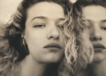 Close-up sepia portrait of model Mila Prvulovic with wavy hair and hoop earring posing next to her mirror reflection.