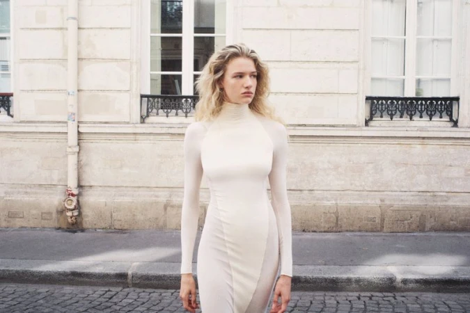 Mila Prvulovic standing outdoors in a fitted cream high-neck dress with long sleeves against a stone building facade.