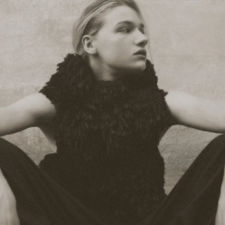 Model Mila Prvulovic sitting in a black textured sleeveless high-neck top and dark trousers against a textured neutral wall.