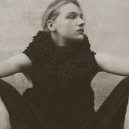 Model Mila Prvulovic sitting in a black textured sleeveless high-neck top and dark trousers against a textured neutral wall.