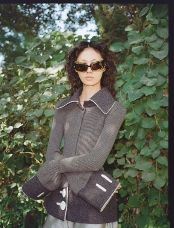 Model Sijia Kang wearing statement sunglasses and a grey ribbed knit jacket with flared cuffs against green foliage.
