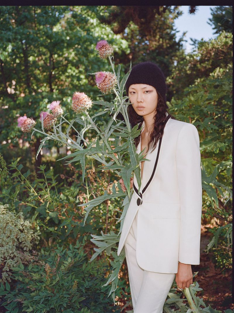 Model Sijia Kang wearing a cream blazer with black trim and a black beanie holding a flowering thistle in a lush green garden.