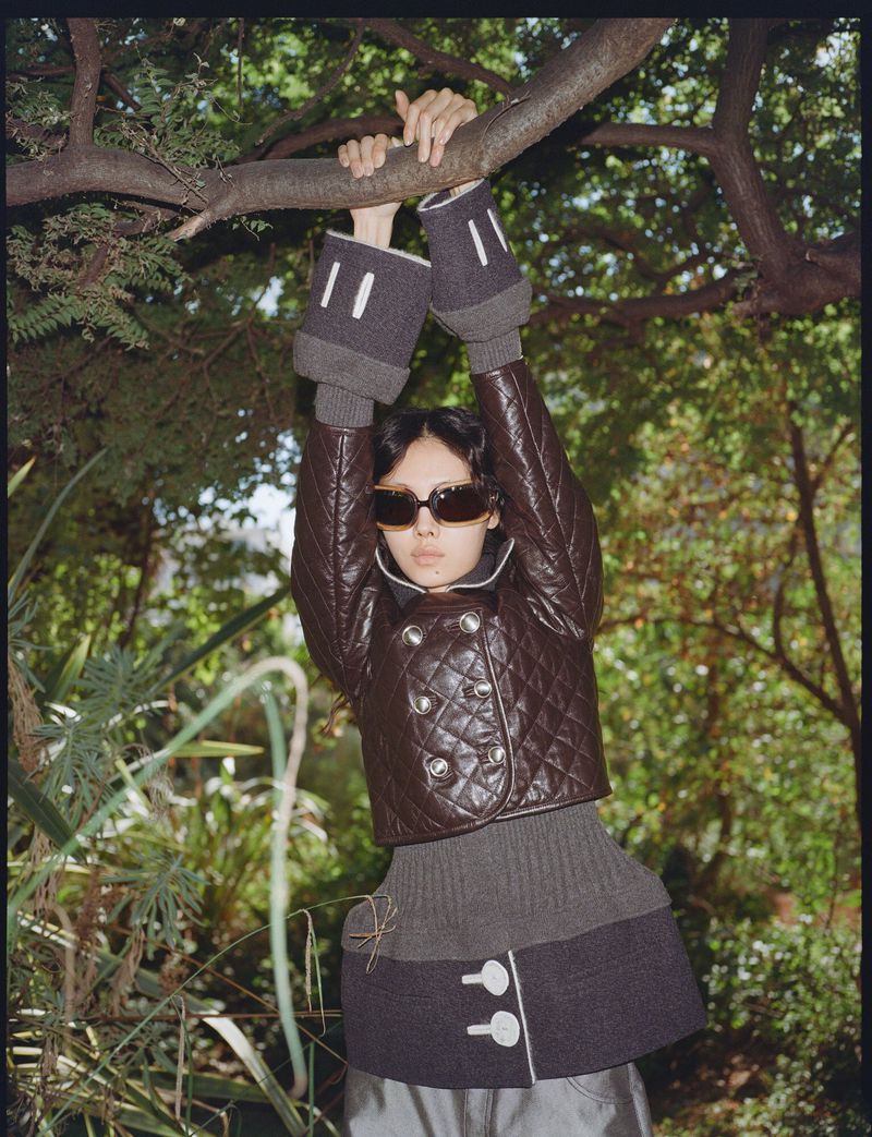 Model Sijia Kang in a brown quilted jacket and sunglasses hanging from a tree branch surrounded by lush green foliage.